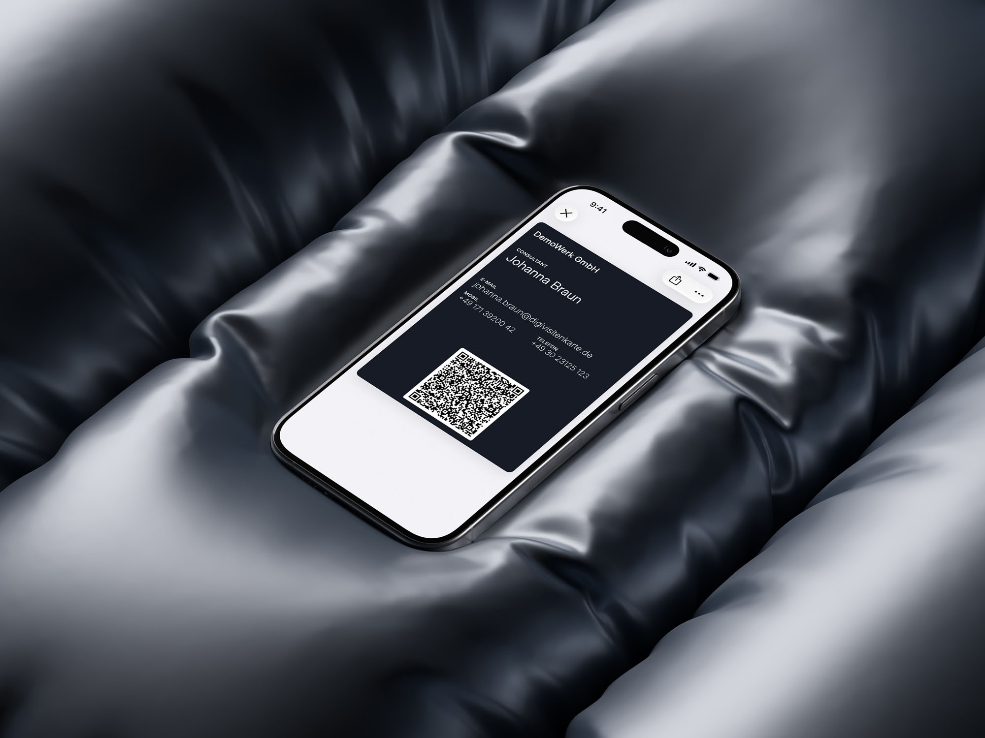 Ein iPhone auf glänzendem schwarzem Stoff zeigt eine dunkelblaue digitale Visitenkarte im Apple Wallet: DemoWerk GmbH, Consultant Johanna Braun, mit E-Mail, Mobilnummer und QR-Code